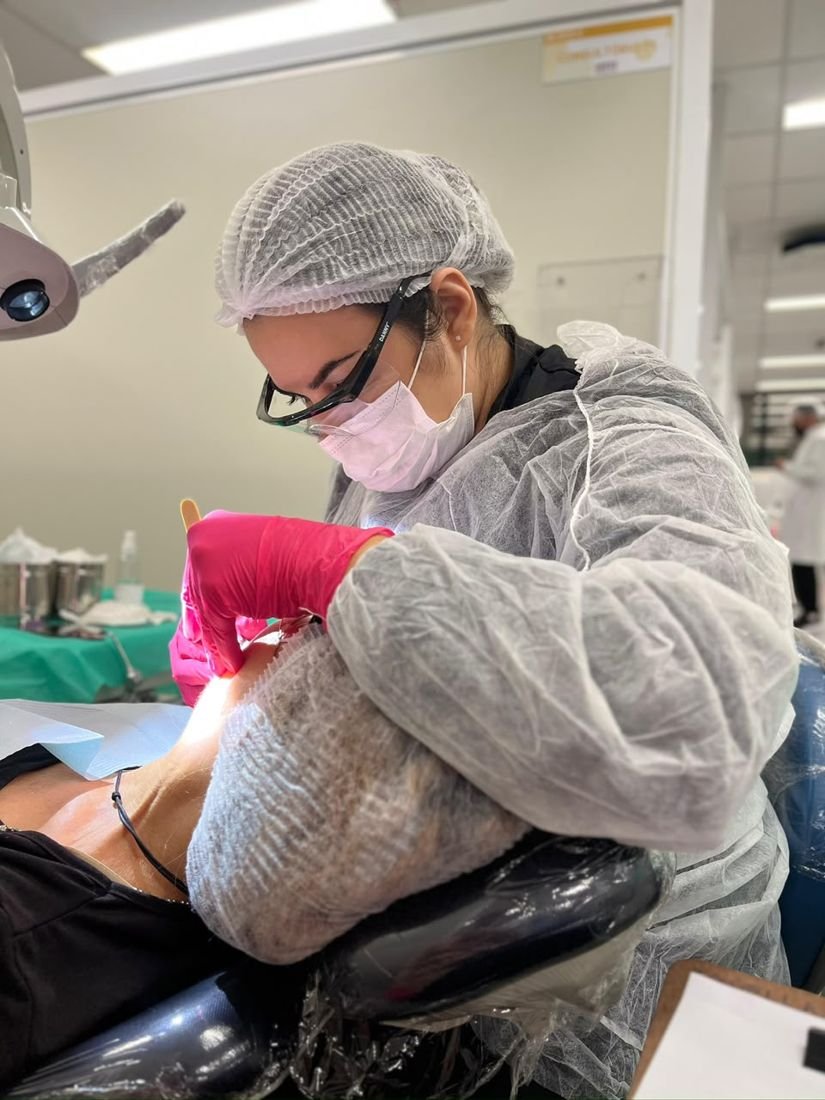 Dra. Kamila atendendo um paciente na cadeira odontológica, com luvas rosa e óculos de proteção.